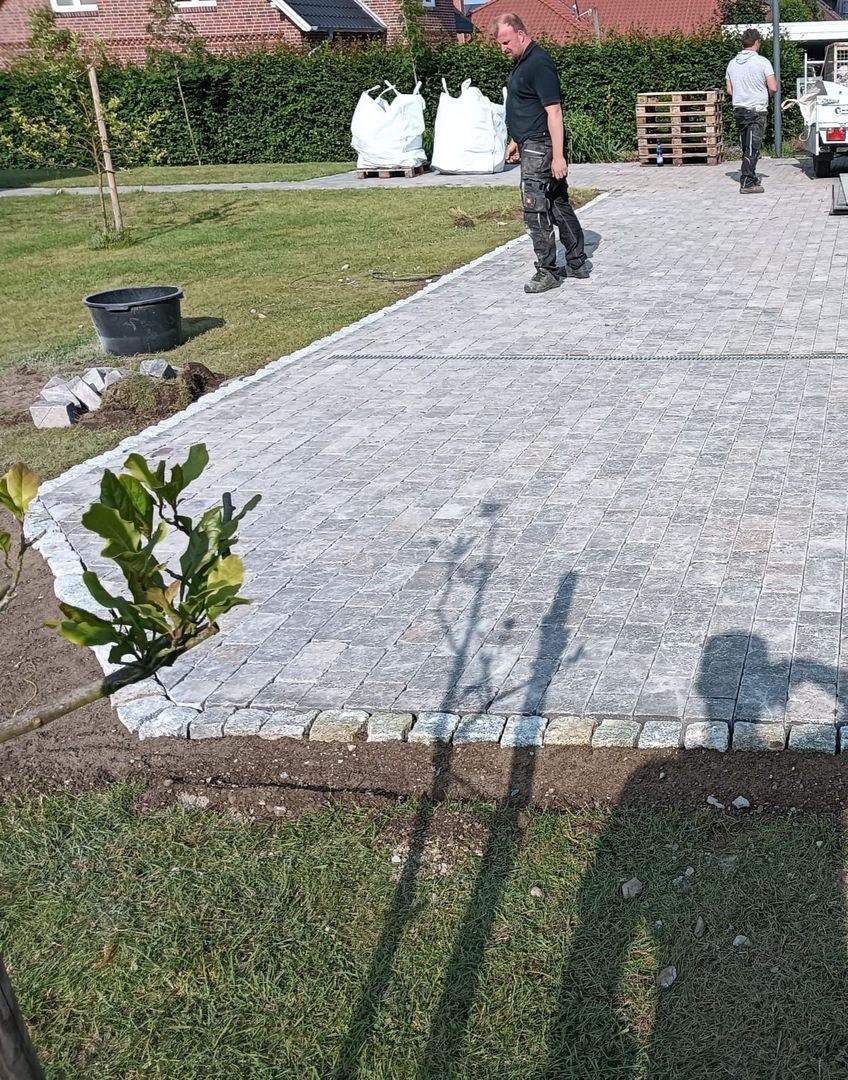 Ein Arbeiter steht auf einem neu gepflasterten Steinweg, umgeben von Baumaterialien und Grünpflanzen, während die Sonne lange Schatten wirft.